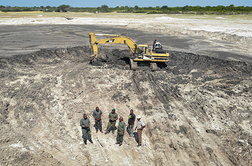 Luchtfoto van zes teamleden in een uitgebaggerd deel van een modderpoel, met het landschap van Hwange National Park op de achtergrond. © IFAW 